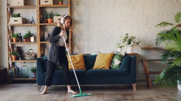 Creative girl is singing and dancing with mop during housework in nice house with beautiful furniture and plants. Fun, domestic work and happy people concept. - Кадры, видео