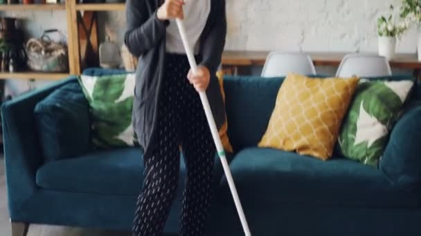 Positive girl is dancing with flat mop and singing doing housework at home, young woman is having fun enjoying music in headphones and washing floor. - Footage, Video