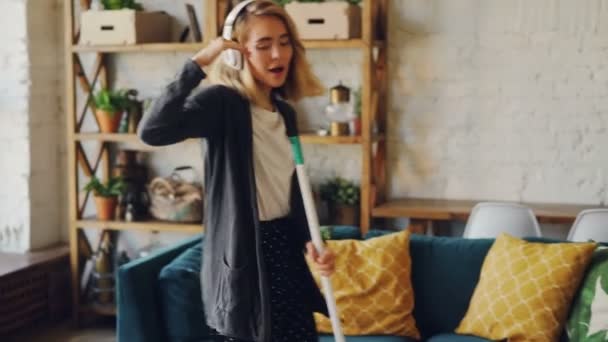 Joyful housewife is holding mop, singing in it and dancing around it enjoying cleanup in loft style apartment. Blond woman is wearing casual clothing and is barefoot. - Filmati, video