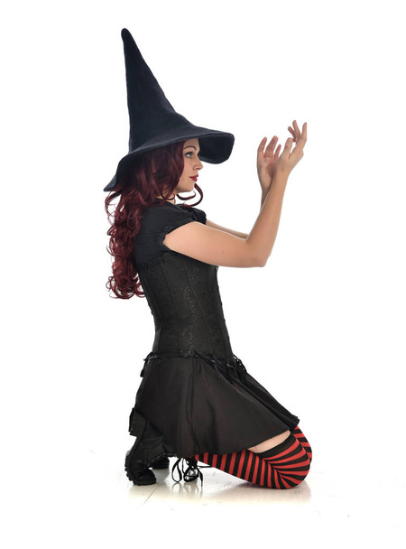 Pełna długość Portret rudowłosy dziewczyna sobie czarny czarownica strój i kapelusz pointy. pozie siedzącej, na białym tle na tle białego studia. - Zdjęcie, obraz
