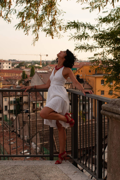 Portrait of a lady on a balcony looking at the city, wearing a white dress, red necklace, wavy dark hair. Happy and very playful and mischievous. Sun at sunset on the city. - Fotoğraf, Görsel