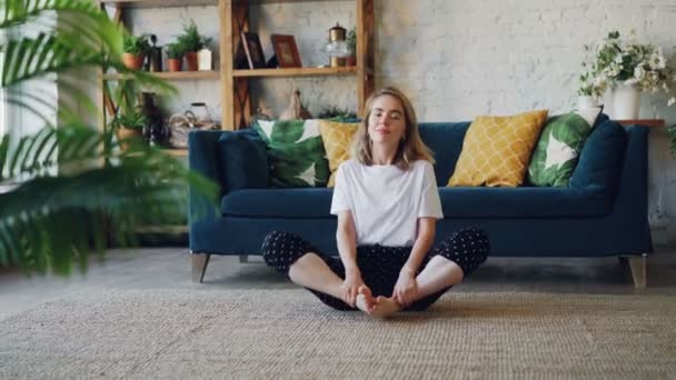 Pretty young woman is practising yoga at home relaxing in simple body position sitting on floor with beautiful furniture around. Healthy lifestyle and people concept. - Video