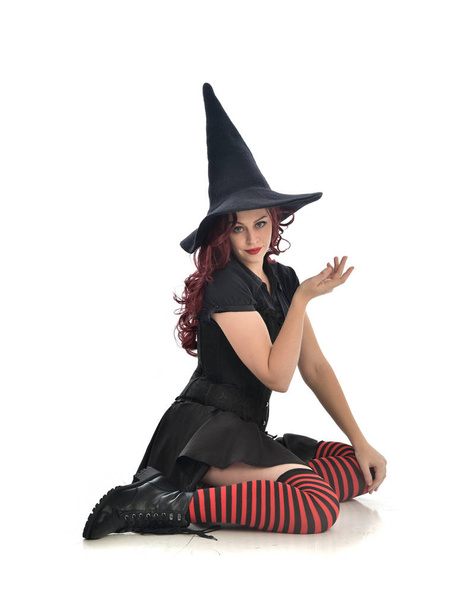retrato de comprimento total da menina de cabelos vermelhos vestindo traje de bruxa preta e chapéu pontiagudo. postura sentada, isolada no fundo do estúdio branco
. - Foto, Imagem