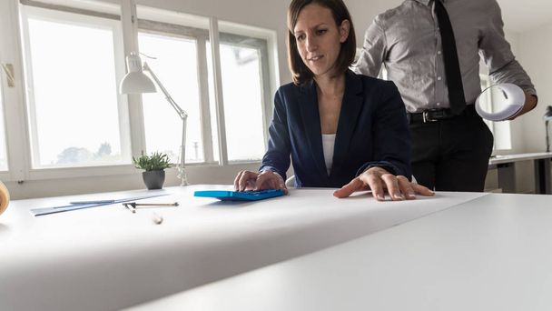Женщина, работающая над синим ручным калькулятором в офисе, в то время как ее партнер стоит позади нее и смотрит на нее под низким углом через стол
. - Фото, изображение