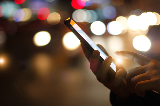 Close-up van vrouwelijke handen mobiele smartphone met verlichte kleur licht bokeh op straat nacht achtergrond - Foto, afbeelding