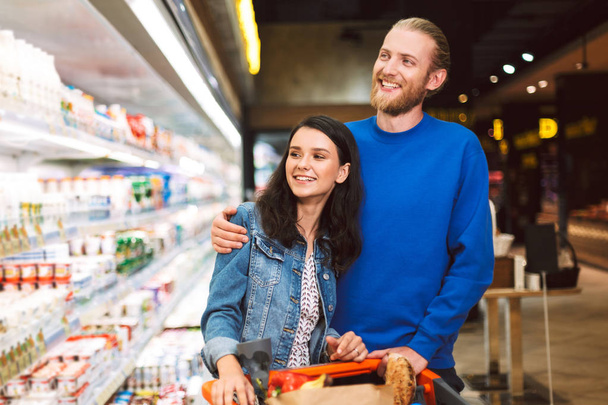 Young cheerful couple with trolley full of products happily choosing dairy in modern supermarket - Photo, Image