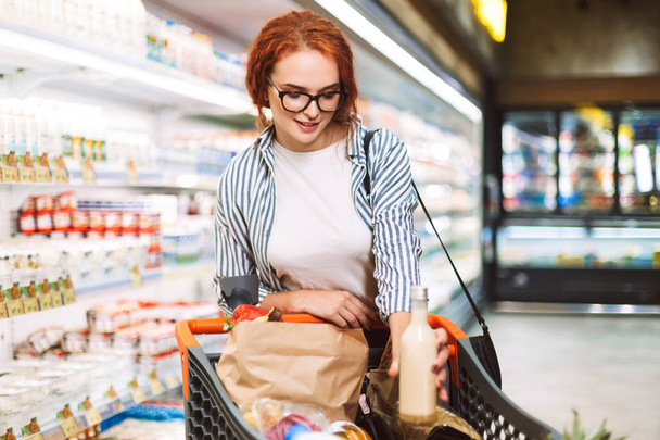 Chica bonita en gafas y camisa a rayas con carrito de compras lleno de productos en el departamento de lácteos de supermercado moderno
 - Foto, imagen