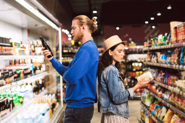 Nuori kaunis pari, joka seisoo selkä selkää vasten harkitusti valitsemalla tuotteita, jotka viettävät aikaa nykyaikaisessa supermarketissa.
  - Valokuva, kuva