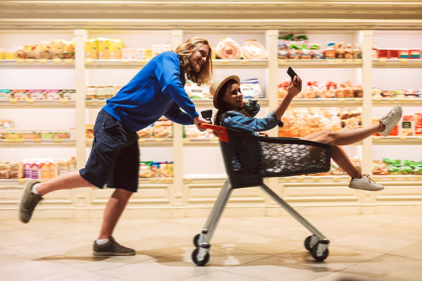 Mladý, usměvavý chlap tlačí nákupního košíku s hezká dívka uvnitř zatímco ona fotí na mobil baví spolu v moderním supermarketu - Fotografie, Obrázek