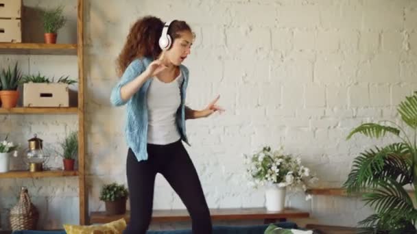 Attractive brunette is having fun at home listening to music through headphones, singing and dancing jumping on sofa. Modern technology and young people concept. - Video