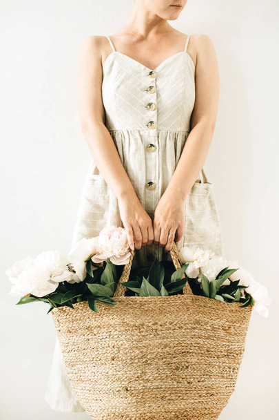 Young pretty woman in linen dress holding straw bag with white peony flowers on white background. - Foto, imagen
