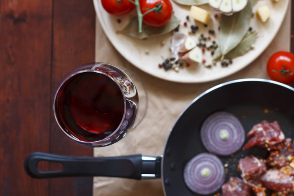 Νωπό βόειο κρέας. έννοια: μαγείρεμα στο σπίτι. Ρηχό βάθος πεδίου. Ένα ποτήρι κρασί, ένα τηγάνι με ωμό κρέας και καρυκεύματα, λαχανικά στο τραπέζι της κουζίνας. Ευρωπαϊκή κουζίνα - Φωτογραφία, εικόνα