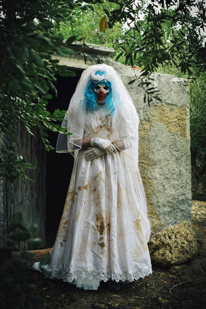 a scary evil clown wearing a dirty and ragged bride dress, at the door of a rustic shelter, in a disturbing rural landscape - Photo, Image