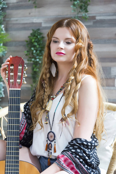 Una hermosa chica con el pelo rizado rojo se sienta en una silla con una guitarra de siete cuerdas. Hippie chica en el estudio
. - Foto, imagen