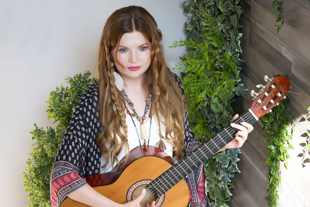 Una bella ragazza con i capelli ricci rossi sta in piedi e tiene una chitarra a sette corde tra le mani. Studio con foglie verdi
. - Foto, immagini