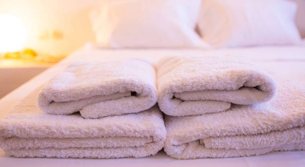 Concepto de habitación de hotel. Primer plano de toallas blancas apiladas en una cama sobre un fondo borroso y cálido iluminado
 - Foto, imagen