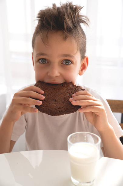 cute boy sitting on kitchen eating chocolate cookie with milk - Foto, imagen