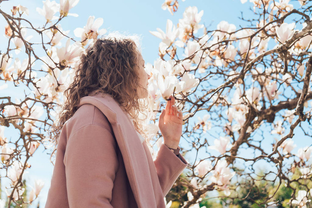 elegant stylish girl enjoys the scent of a blooming pink magnolia in a park in the rays of the spring sun - Фото, изображение