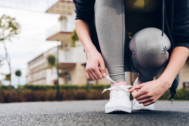 струнка дівчина в сірих спортивних штанях зв'язує свої черевики на кросівки, готується до бігу
 - Фото, зображення