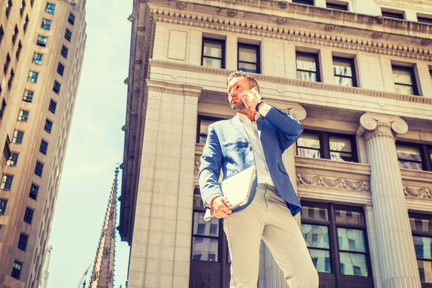 Jeune homme d'affaires européen avec barbe voyageant, travaillant à New York, portant un blazer bleu, un pantalon gris, tenant un ordinateur portable, se tenant devant un vieil immeuble de bureaux, appelant sur un téléphone portable - Photo, image