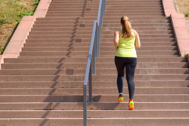 Visão traseira de comprimento total de uma mulher ativa e determinada de meia-idade correndo enquanto subia escadas durante o treino intenso para perda de peso ao ar livre em um dia ensolarado
 - Foto, Imagem