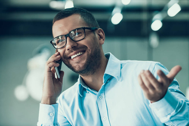 Young Smiling Businessman Talking on Phone. Happy Man wearing Glases and blue Shirt Talking on Smartphone at Workplace. Man enjoying Work at Office. Corporate Lifestyle Concept - 写真・画像