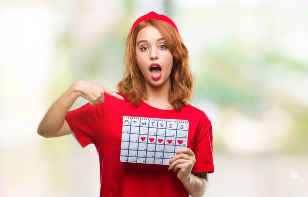 Молодая красивая женщина на изолированном фоне держит календарь менструации с удивленным лицом указывая пальцем на себя
 - Фото, изображение