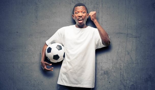 African black man holding soccer ball happy and excited celebrating victory expressing big success, power, energy and positive emotions. Celebrates new job joyful - Foto, Imagem