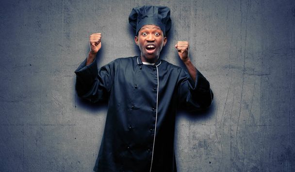 Jonge zwarte koken dragen chef-kok hoed blij en opgewonden vieren overwinning groot succes, kracht, energie en positieve emoties te uiten. Nieuwe baan vreugdevolle viert - Foto, afbeelding