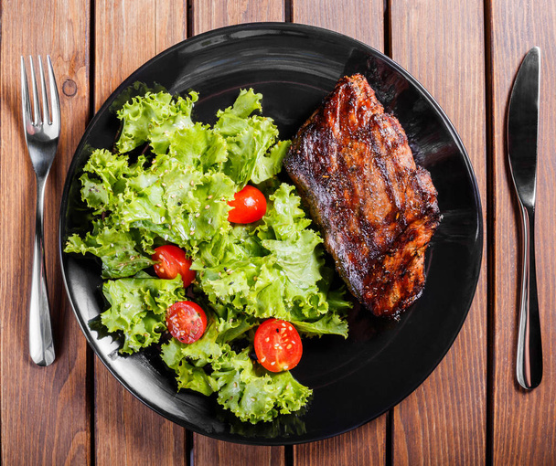 Grilled pork steak meat with fresh vegetable salad and tomatoes on black plate, wooden background. - Photo, Image