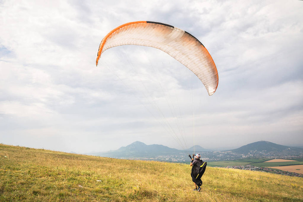 A man paraglider taking off from the edge of the mountain with fields in the background. Paragliding sports - Фото, изображение