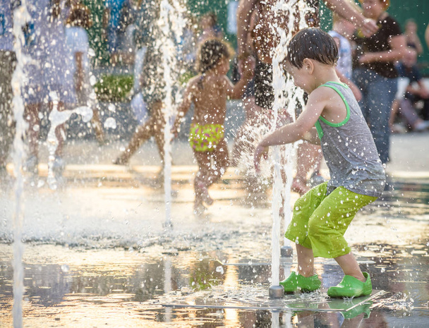 Opgewonden jongen plezier tussen waterstralen, in de fontein. Zomer in de stad. Het gezicht van de tevreden glimlach van het kind. Ecologie concept. - Foto, afbeelding