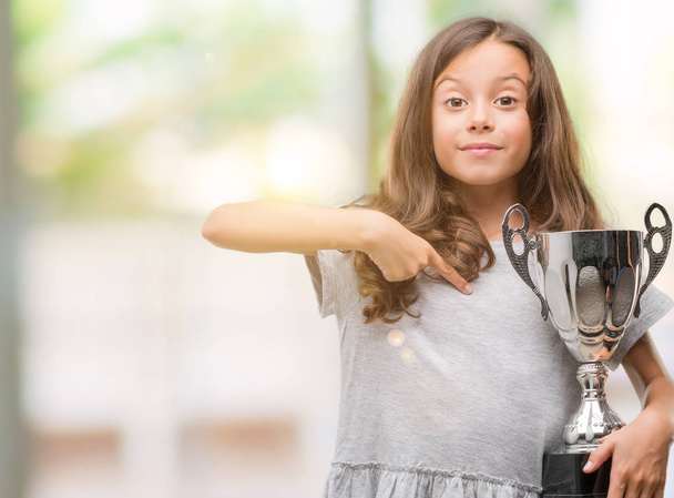 Brunette hispanic girl holding a trophy with surprise face pointing finger to himself - Photo, Image