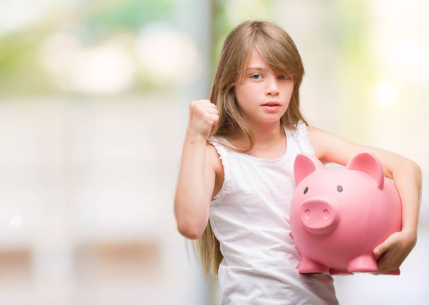 Young blonde toddler holding piggy bank annoyed and frustrated shouting with anger, crazy and yelling with raised hand, anger concept - Photo, Image