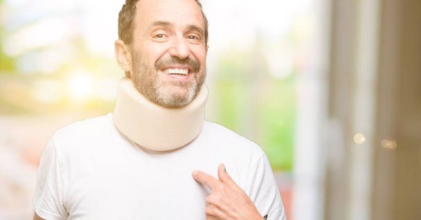 Injured senior man using a neck brace happy and surprised cheering expressing wow gesture, pointing with finger - 写真・画像