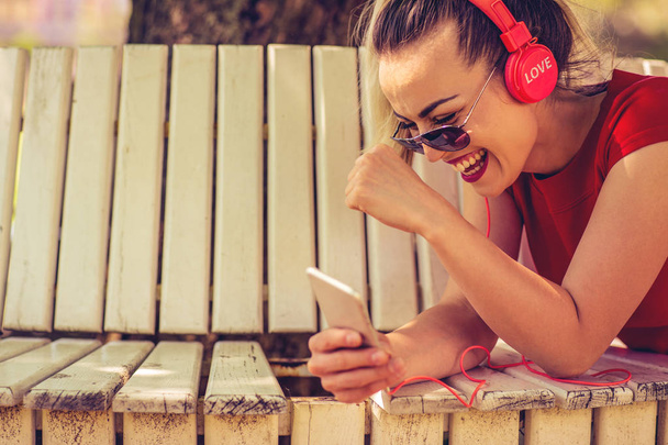 Jovem feliz ouvindo a música de um smartphone deitado em um banco de um parque de perto
 - Foto, Imagem