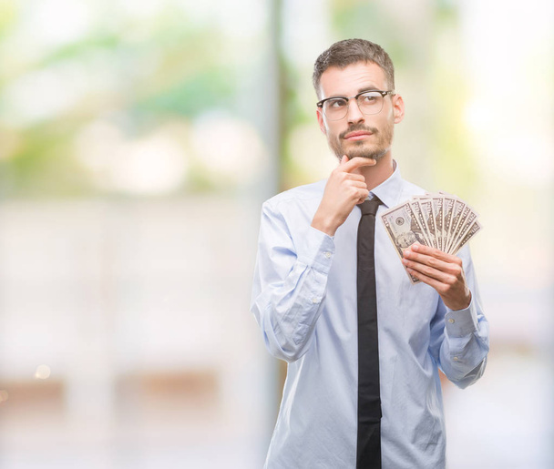 Joven hombre de negocios hipster sosteniendo dólares cara seria pensando en la pregunta, idea muy confusa
 - Foto, Imagen