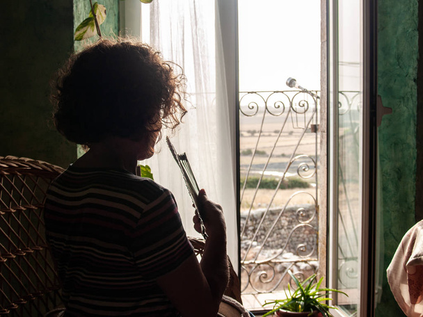 Portrait de femme mature avec des cheveux bouclés et des lunettes et ventilateur espagnol dans une maison rurale regardant par une fenêtre ouverte
 - Photo, image