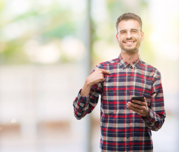 Young hipster adult man holding smartphone with surprise face pointing finger to himself - Photo, Image