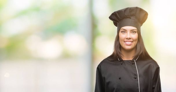 Jeune cuisinière hispanique portant l'uniforme de chef avec un visage heureux debout et souriant avec un sourire confiant montrant les dents
 - Photo, image