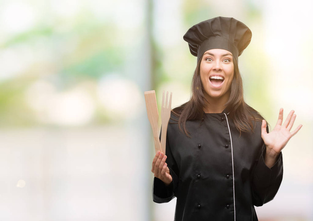 Young hispanic cook woman wearing chef uniform very happy and excited, winner expression celebrating victory screaming with big smile and raised hands - Photo, Image