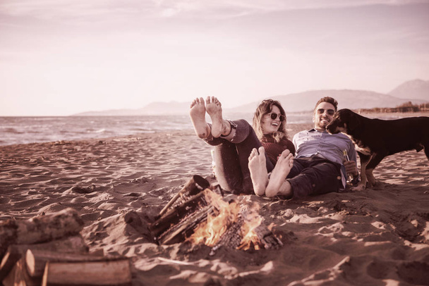 Young Couple Relaxing By The Fire, Drinking A Beer Or A Drink From The Bottle on the beach at autumn day filter - Фото, изображение