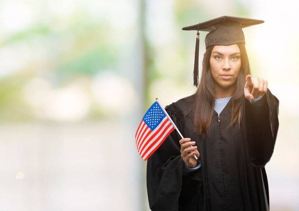 Jovem hispânica vestindo uniforme graduado segurando bandeira da América apontando com o dedo para a câmera e para você, sinal de mão, gesto positivo e confiante da frente
 - Foto, Imagem