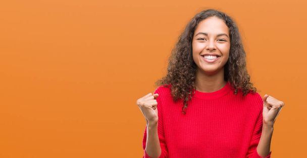 Young hispanic woman wearing red sweater celebrating surprised and amazed for success with arms raised and open eyes. Winner concept. - Foto, imagen