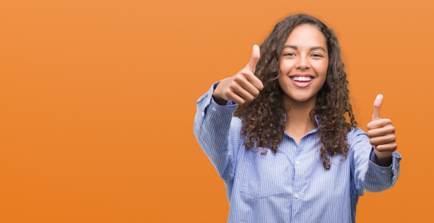 Young hispanic business woman approving doing positive gesture with hand, thumbs up smiling and happy for success. Looking at the camera, winner gesture. - Photo, Image