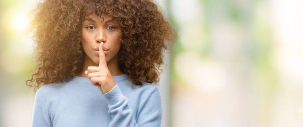Donna afroamericana con un maglione che chiede di stare zitta con un dito sulle labbra. Silenzio e concetto segreto
. - Foto, immagini