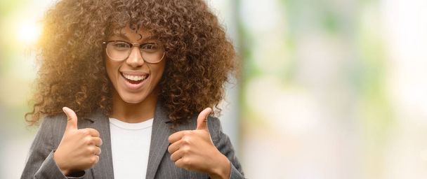 African american business woman wearing glasses success sign doing positive gesture with hand, thumbs up smiling and happy. Looking at the camera with cheerful expression, winner gesture. - Photo, Image
