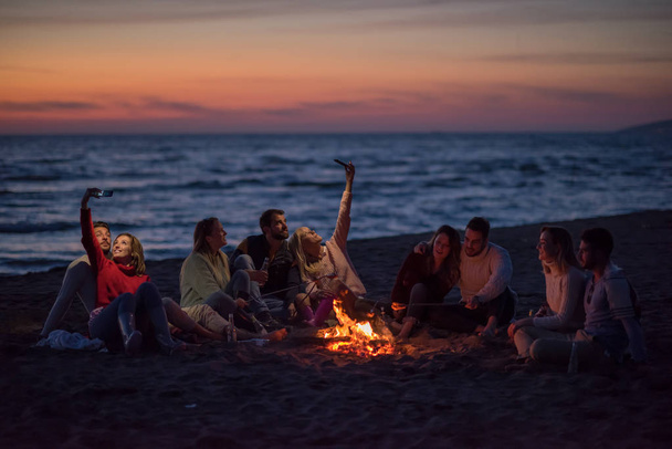 a group of friends using cell phones around the bonfire during autumn beach party drinking beer and having fun - Foto, immagini