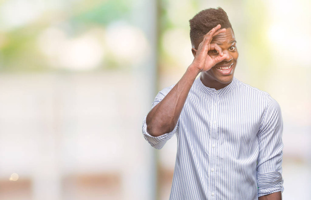 junger afrikanisch-amerikanischer Mann über isoliertem Hintergrund macht ok Geste mit lächelnder Hand, Auge blickt durch Finger mit glücklichem Gesicht. - Foto, Bild