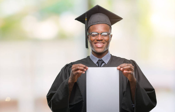Jovem graduado americano africano segurando papel branco grau sobre fundo isolado com um rosto feliz de pé e sorrindo com um sorriso confiante mostrando dentes
 - Foto, Imagem